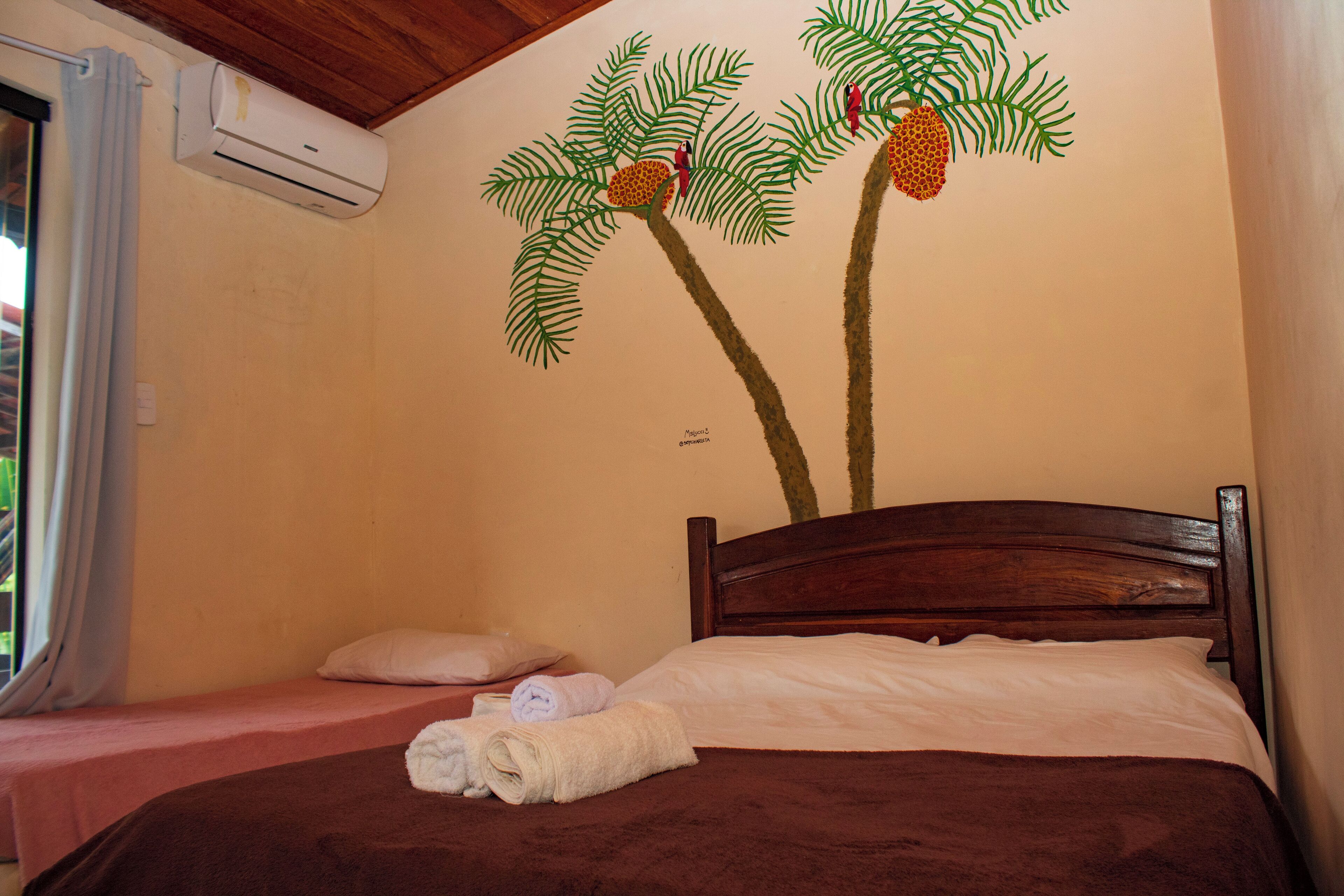 A bedroom with a wooden headboard, two beds, a wall mural of palm trees, and an air conditioning unit.