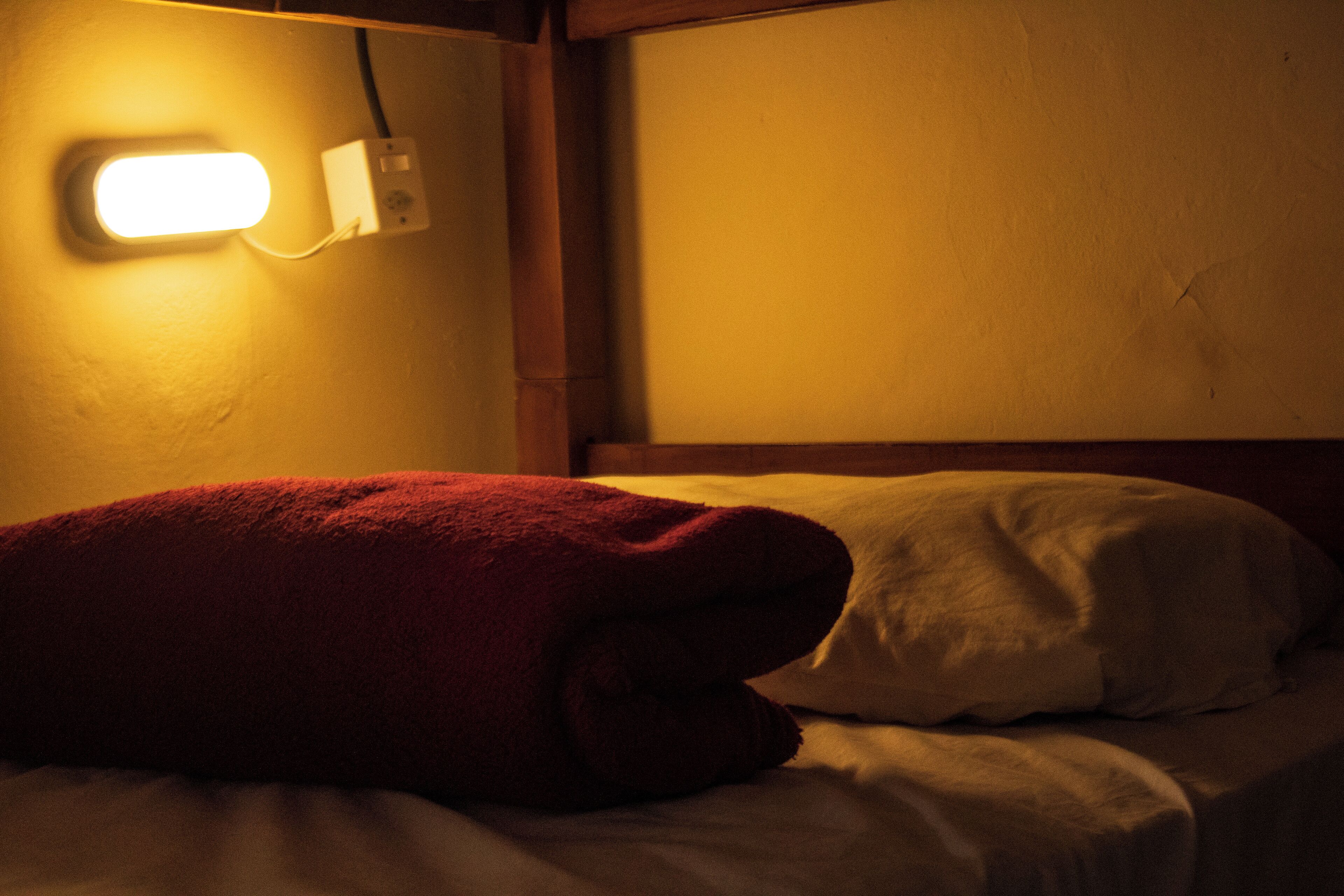 Cama com manta vermelha e almofadas brancas, uma tomada e cabeceira de madeira.