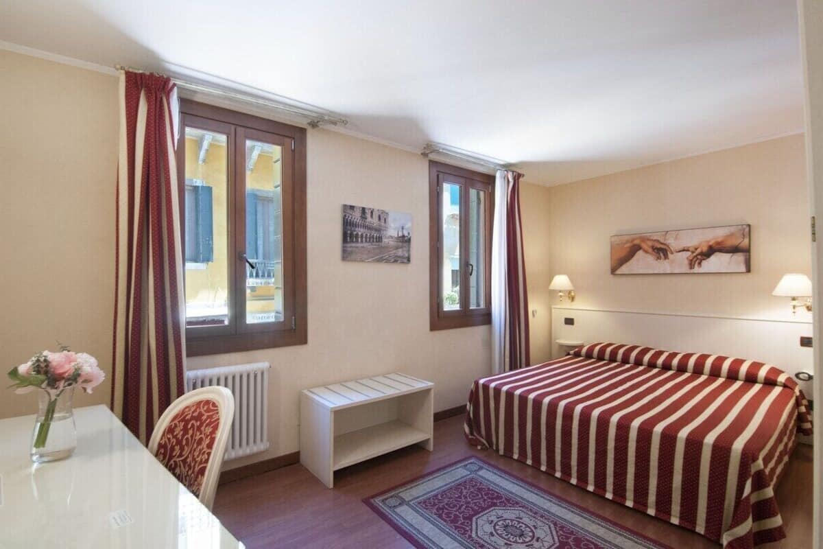 A hotel room with a bed, a small table with a vase of flowers, a chair, a radiator, and a window with curtains.
