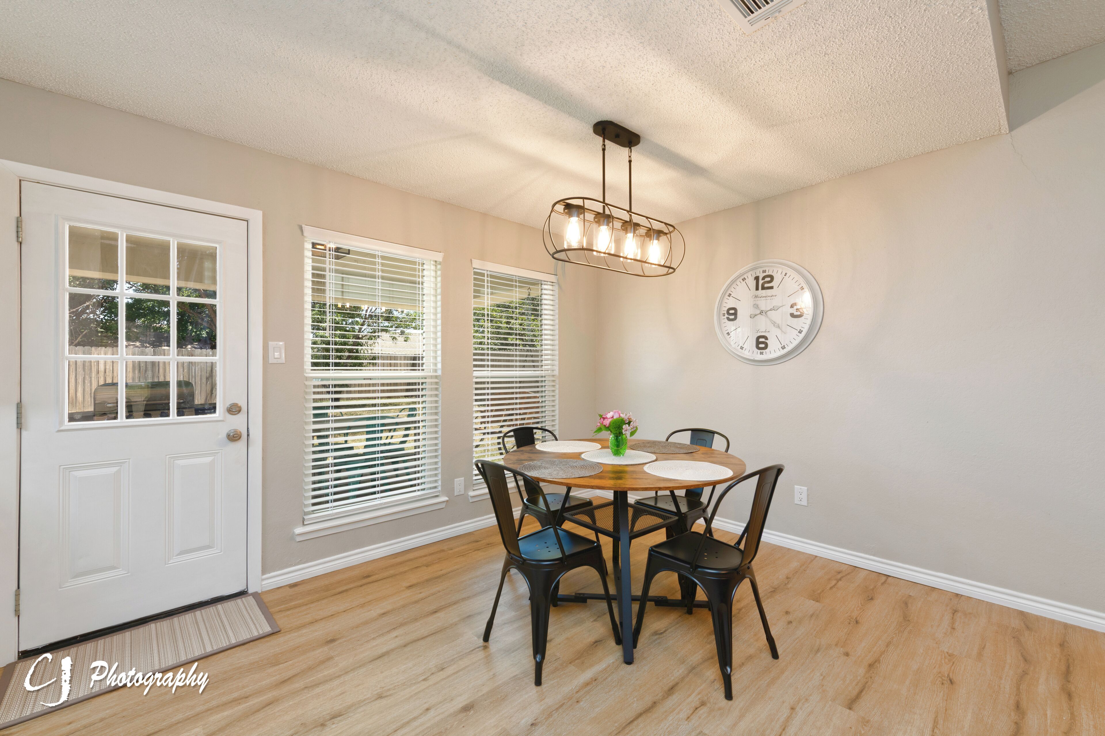 A dining area with a round table, four chairs, a chandelier, a clock, and a door with glass panels.