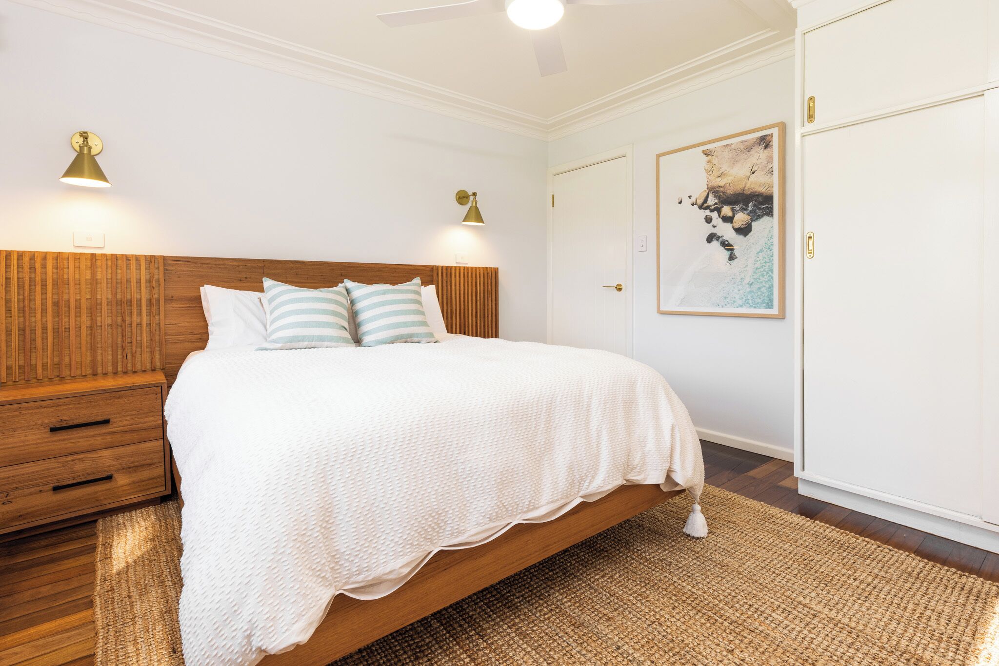 Une chambre à coucher avec une tête de lit en bois, un couvre-lit blanc, deux lampes fixées au mur, une œuvre d’art encadrée sur le thème de la côte et une grande armoire.