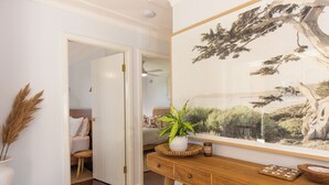 A room with a wooden floor, a white door leading to another room, a wooden console table with a plant, a framed picture, and a small wooden side table with a candle. - Cove Cottage South West Rocks (South West Rocks)