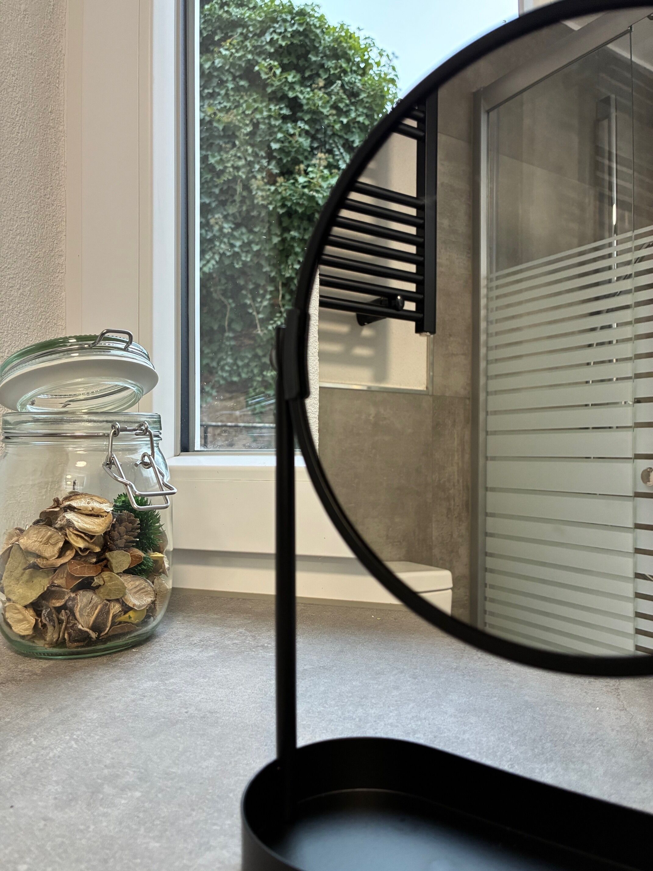 Una encimera de cocina con un tarro de vidrio que contiene hojas secas y piñas, un espejo con marco negro y una ventana con persianas.