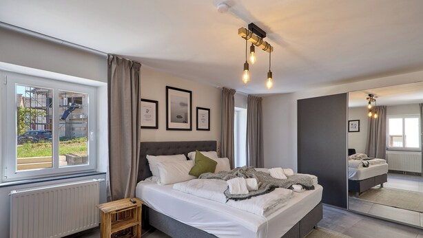 A modern bedroom with a large bed, a bedside table, a mirror, and a window with a view of the outside.