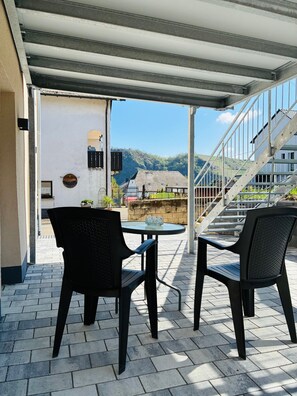 A covered outdoor seating area with two chairs and a table, a staircase, and a view of a mountain and buildings in the background. - Moselquartier "Reben Apartment" up to 4 persons `modern kitchen` box spring bed (Piesport)