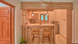 A compact kitchen with a wooden bar counter, two bar stools, pendant lights, and a window with a green frame. - Sarang Resort Varkala by VOYE HOMES (Varkala)