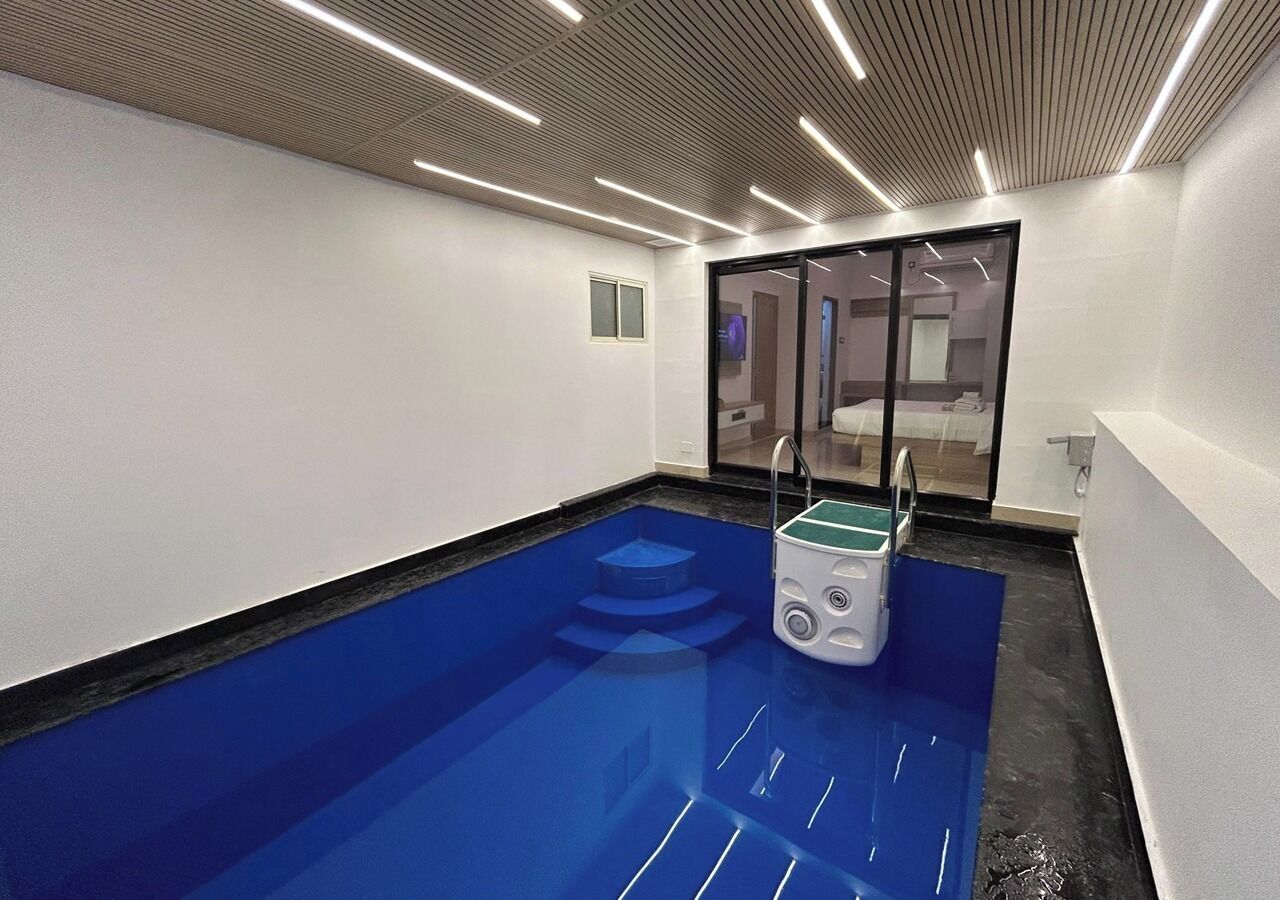 A modern indoor swimming pool with a blue floor, a small pool lift, and a glass door leading to a changing room.