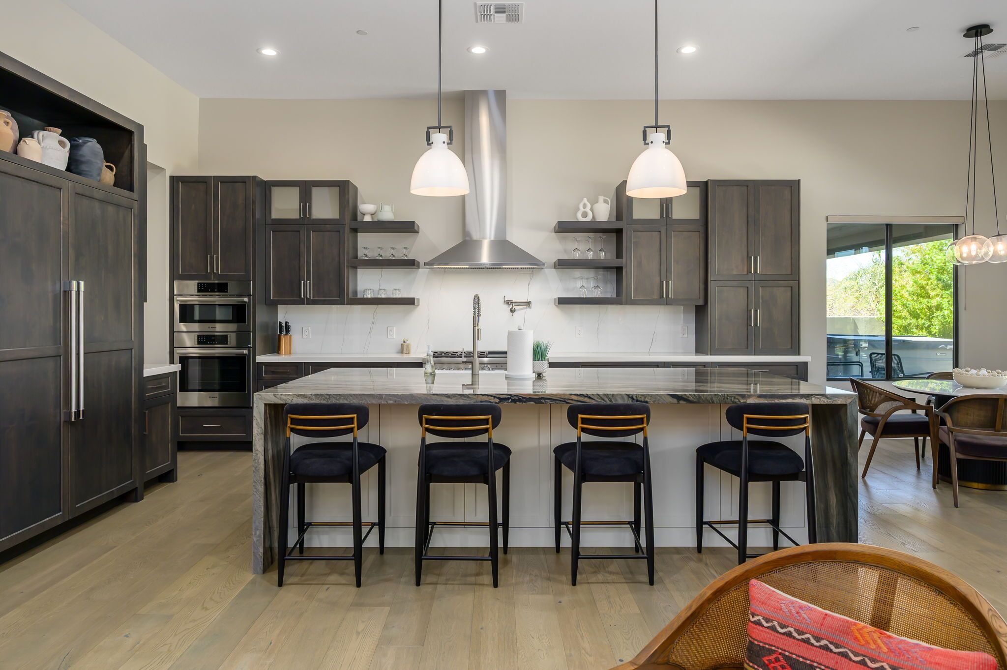 Una cocina moderna con armarios oscuros, electrodomésticos de acero inoxidable, una isla central con taburetes altos, iluminación colgante y un ventanal grande.