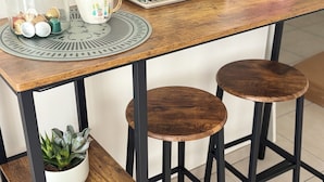 A kitchen with a wooden table, two stools, a coffee maker, a mug rack, and a glass jar with utensils.