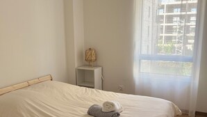 A neatly made bed with a white and gray towel, a small bedside table with a lamp, and a window with a view of buildings.