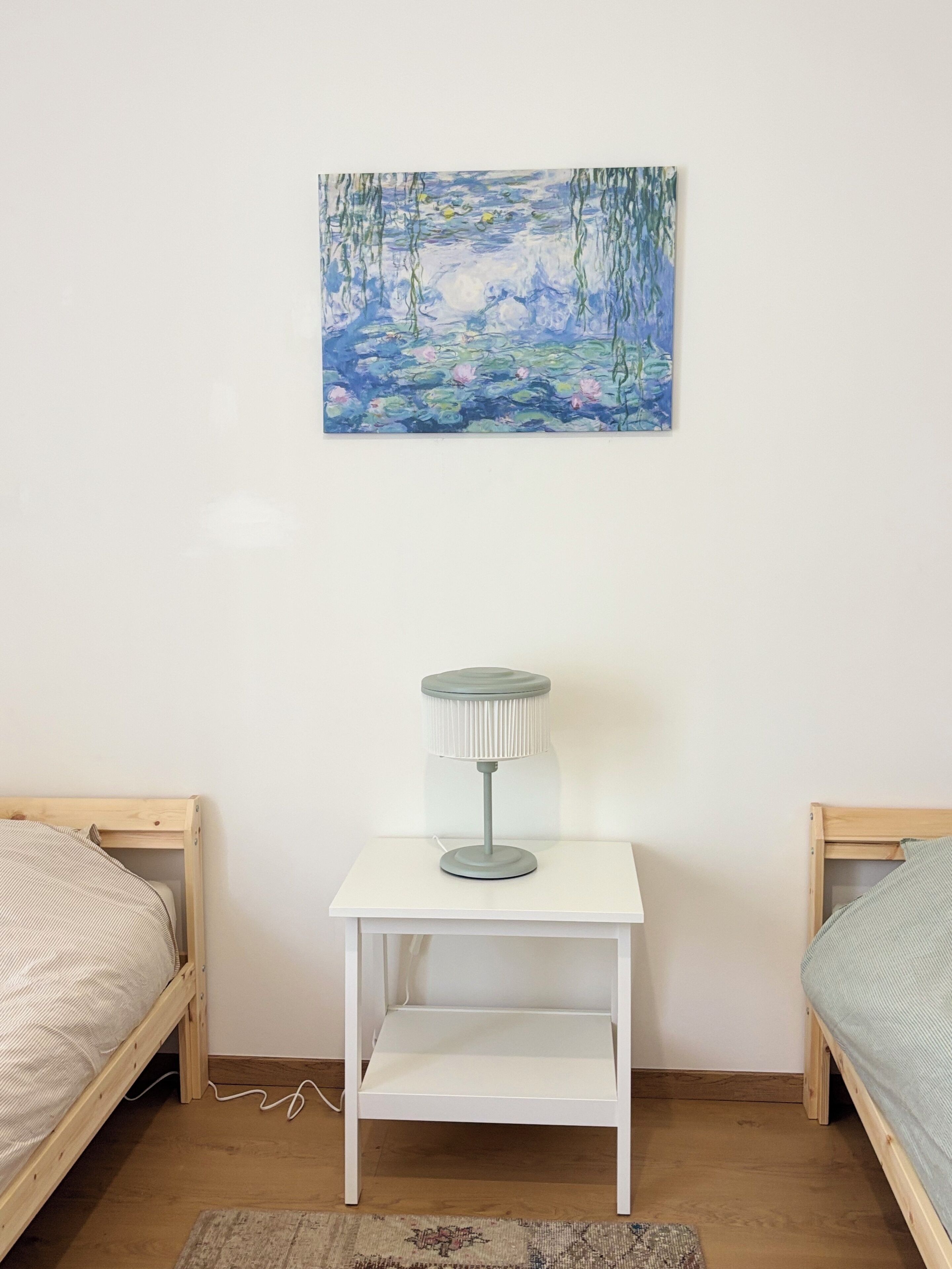 Un dormitorio con una mesita de noche blanca, una lámpara y un cuadro de un paisaje azul.