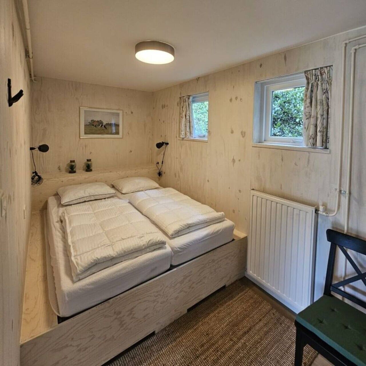A room with a double bed, a small table, a chair, a window with curtains, a picture on the wall, and a radiator.