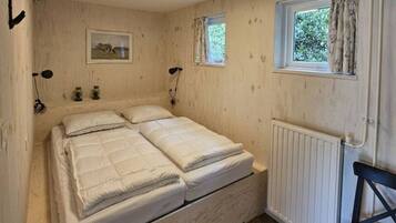A room with a double bed, a small table, a chair, a window with curtains, a picture on the wall, and a radiator.