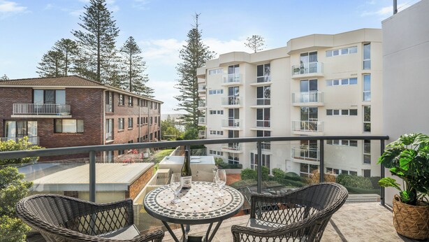 A balcony with wicker chairs and a table, overlooking a residential area with apartment buildings and trees.