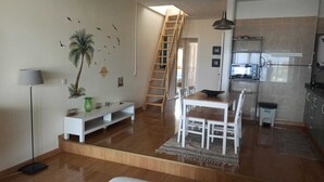 Un salon moderne avec un plancher en bois, une table à manger blanche avec des chaises, un canapé blanc, un petit meuble blanc, un vase vert et un escalier menant à une chambre à coucher située en mezzanine.