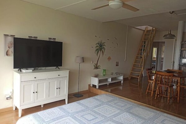 Amplia sala de estar con un televisor de pantalla plana grande sobre un mueble blanco, una lámpara de pie, una pequeña mesa con una planta, una silla de madera y una escalera que lleva a otra habitación.