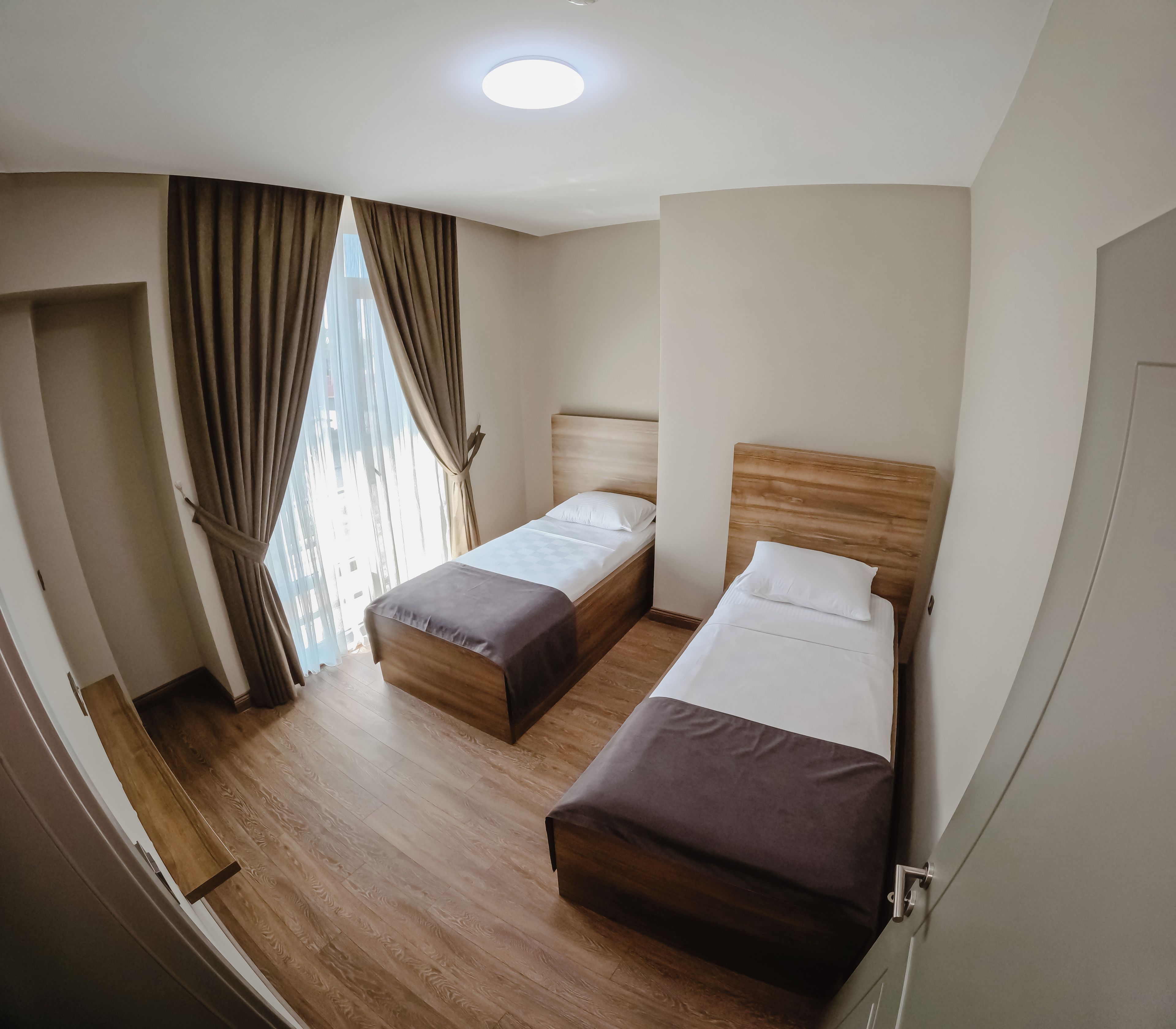 A hotel room with two beds, wooden flooring, a window with curtains, and a ceiling light.