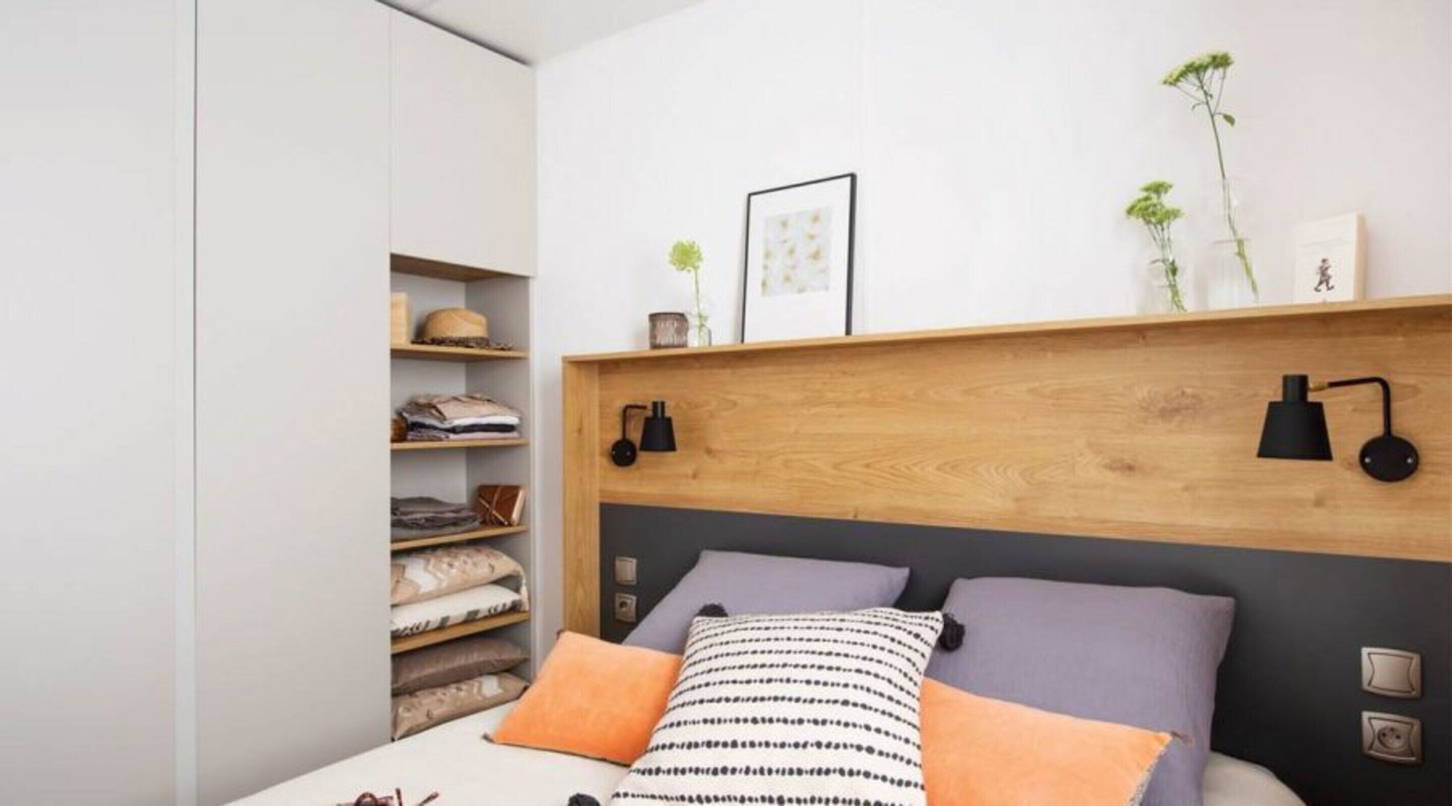 A bedroom with a bed, a wooden headboard, a shelf with folded clothes, a framed picture, and a small plant.