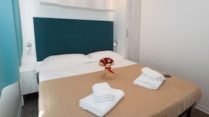 A hotel room with a bed, a nightstand, a wall-mounted shelf, and a bouquet of flowers on the bed. - The Garda Village Caravan Park, Sirmione (Sirmione)