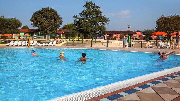 A swimming pool with lounge chairs and umbrellas, surrounded by trees and buildings. - The Garda Village Caravan Park, Sirmione (Sirmione)