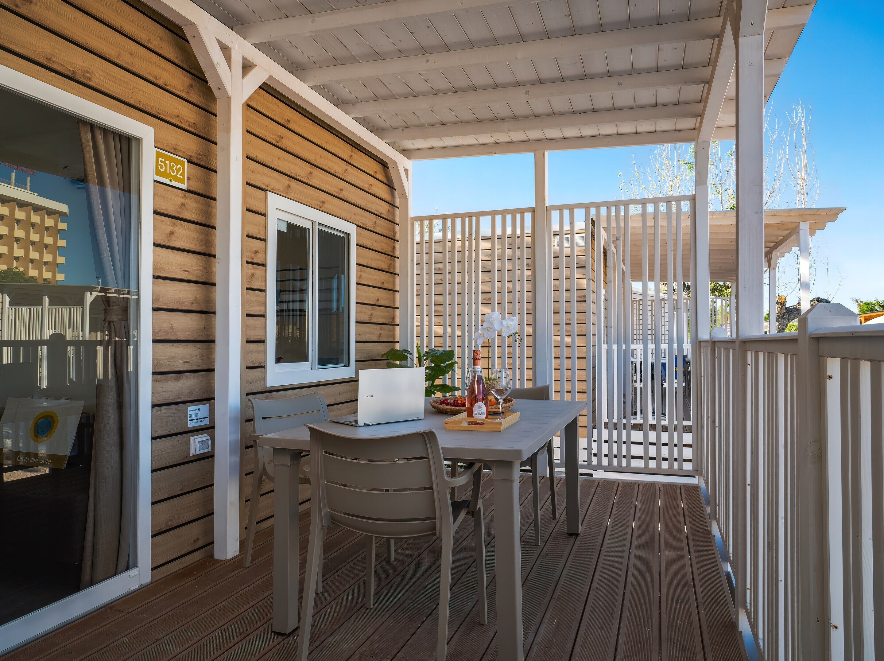 A wooden deck with a table and chairs, a laptop, a bottle of wine, and a glass on the table.
