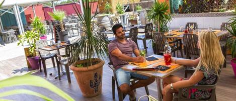 Un café al aire libre con mesas y sillas de madera, plantas en macetas y una estructura de techo de paja al fondo.