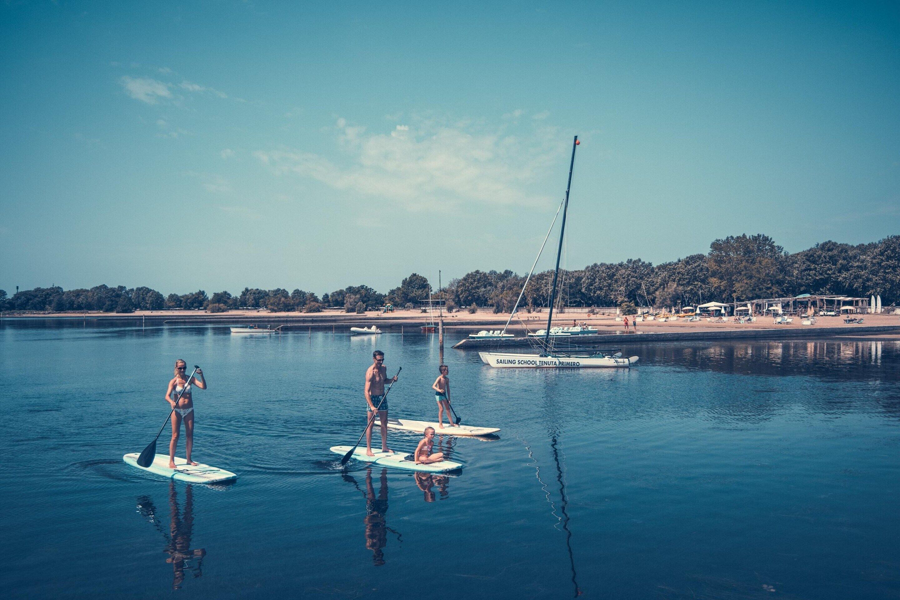 Ein ruhiger See mit Stand-Up-Paddles, einem Segelboot und einem Strand mit Sonnenschirmen im Hintergrund.
