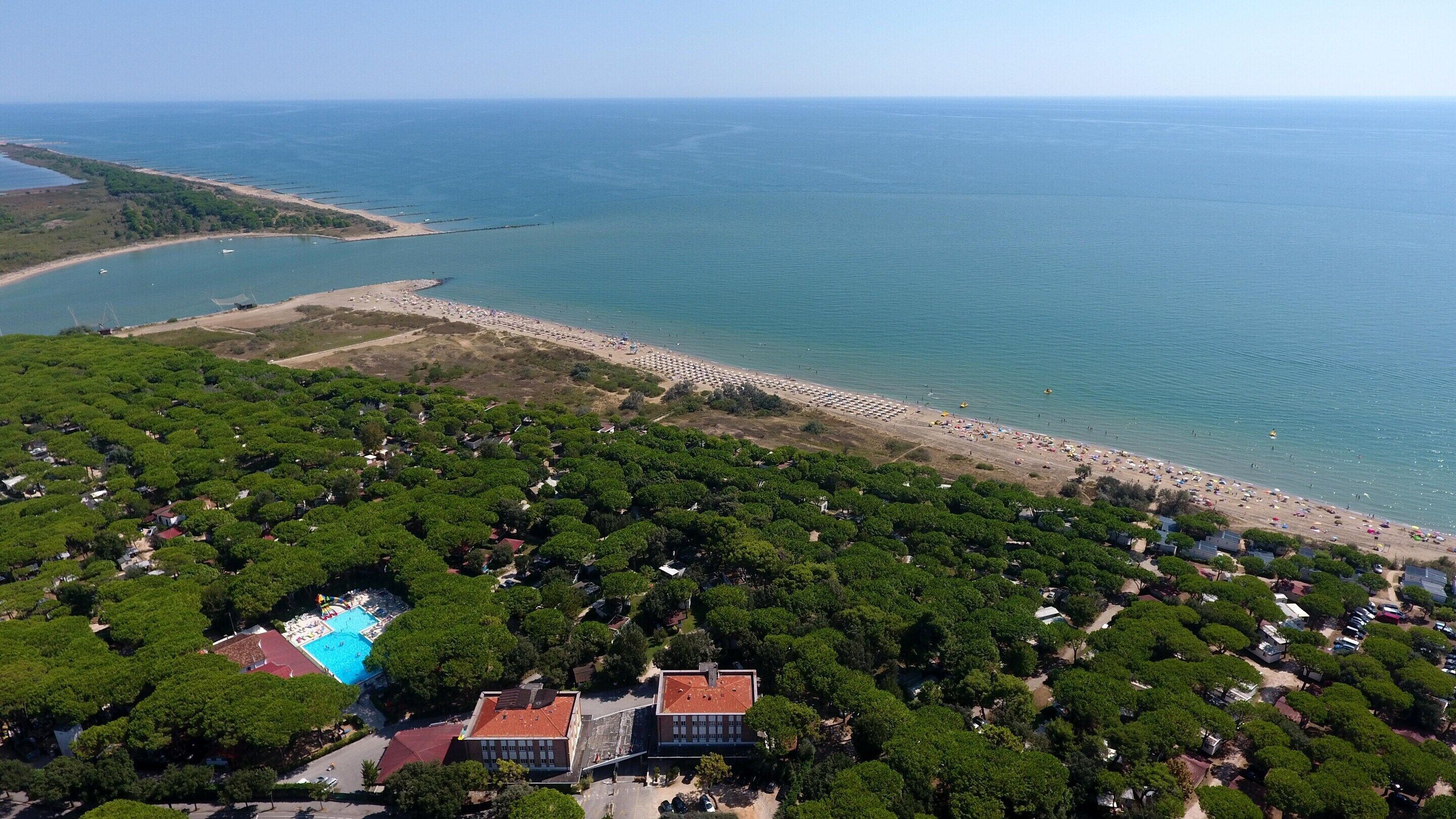 An aerial view of a coastal area with a beach, a swimming pool, and dense greenery.