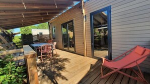 A wooden deck with a table and chairs, a sliding glass door, and a red lounge chair. - Mobile Home Prestige With air Conditioning (Sérignan)