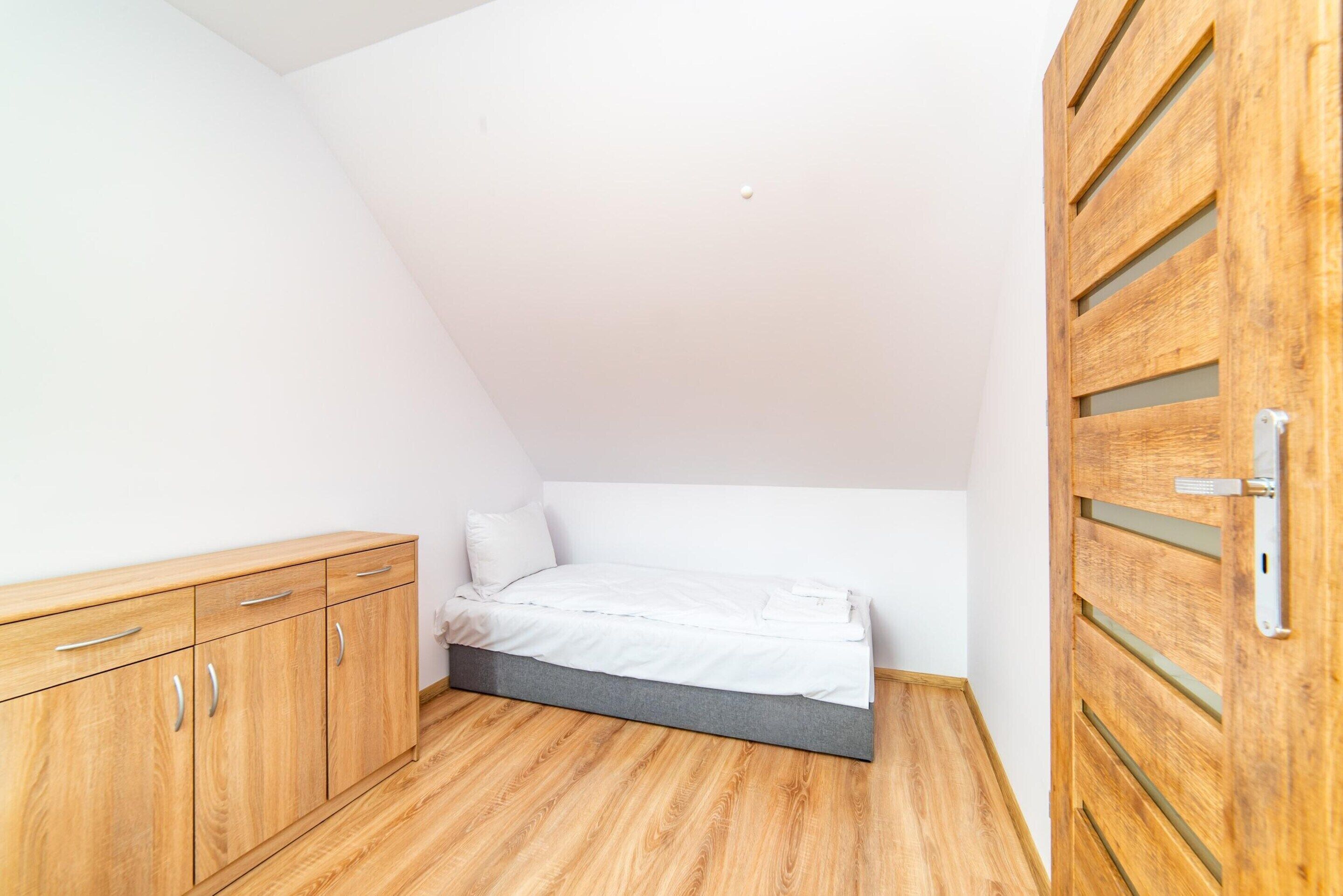 Una habitación pequeña con cama individual, techo inclinado, armario de madera y puerta de madera.