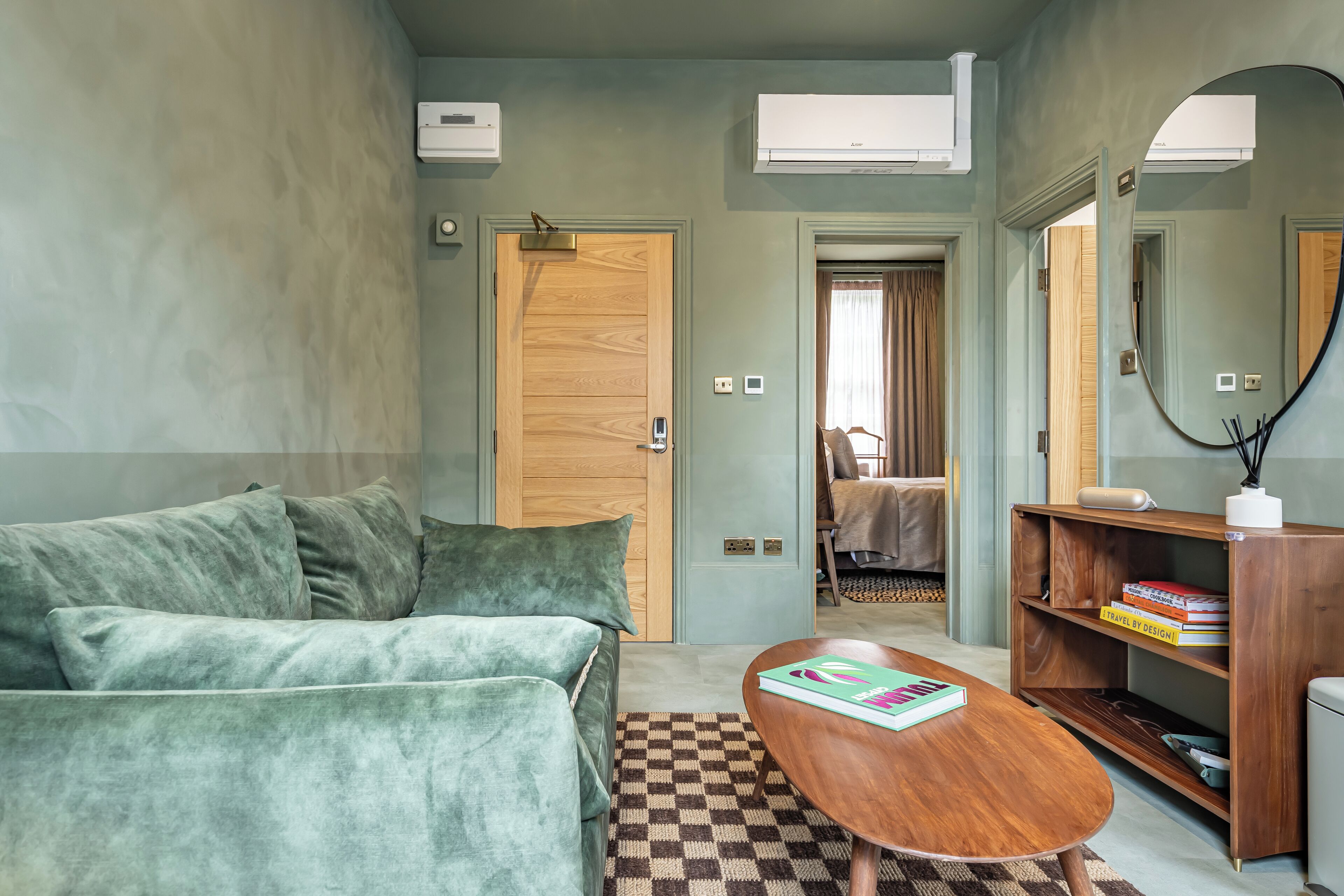 A compact living space with a green sofa, a wooden coffee table, and a checkered rug. There is a bookshelf with books and a mirror.
