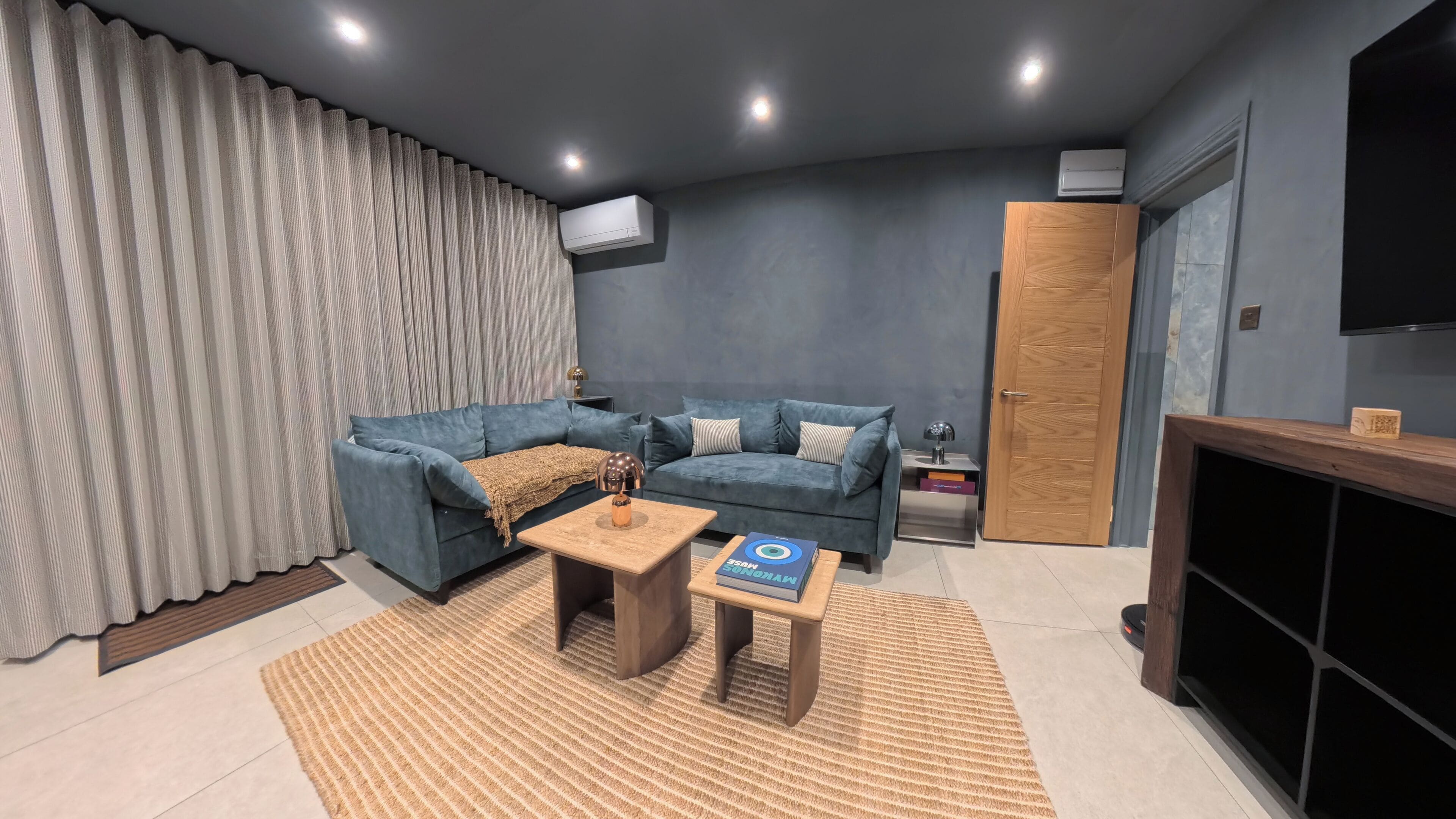 Uma sala de estar moderna com um sofá seccional azul, uma pequena mesa de centro de madeira e uma TV fixada na parede.