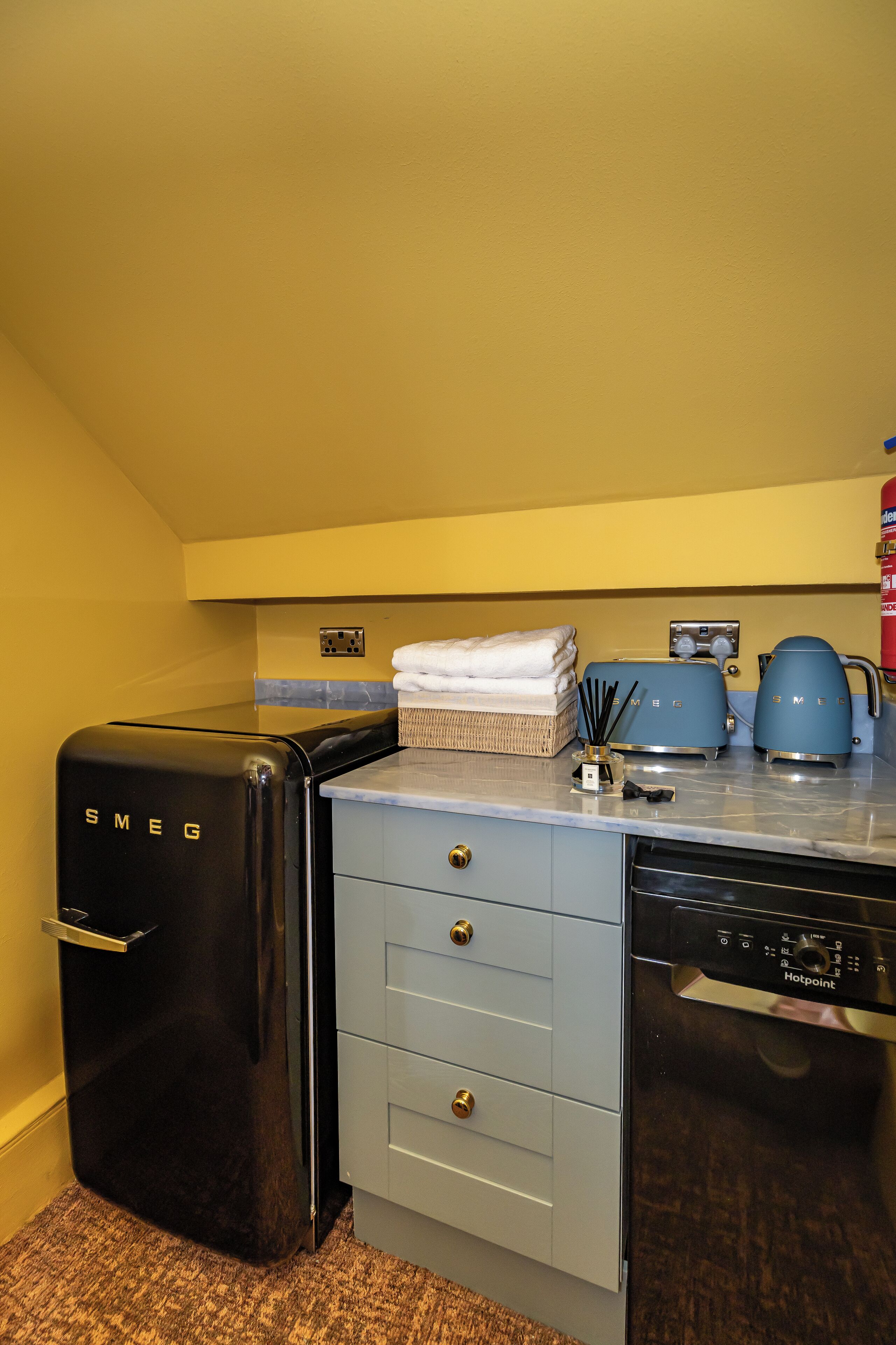 A compact kitchenette with a black Smeg refrigerator, a blue toaster, a coffee maker, and a dishwasher.
