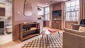 Una sala de estar moderna con un sofá, una mesa de centro y una estantería. Hay una zona de cocina con microondas y un televisor. La habitación tiene ventanas grandes con cortinas.