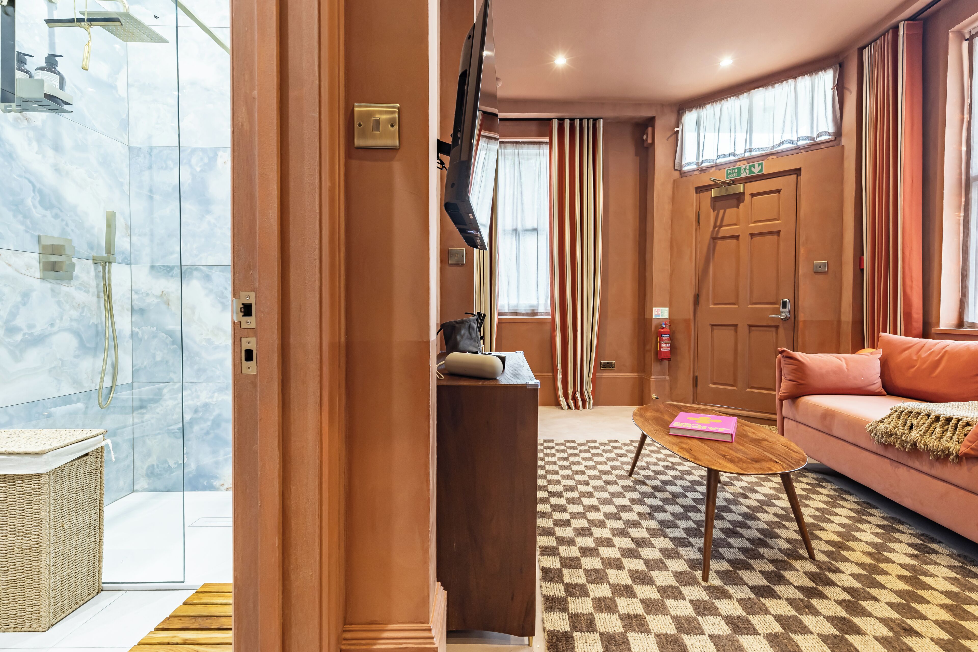 Habitació d'hotel amb un paviment a escacs, un escriptori de fusta, un sofà rosa, un televisor, una zona de dutxa i una porta amb senyal d'emergència.
