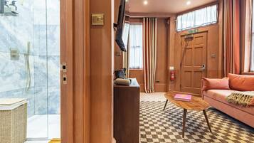 Habitació d'hotel amb un paviment a escacs, un escriptori de fusta, un sofà rosa, un televisor, una zona de dutxa i una porta amb senyal d'emergència.