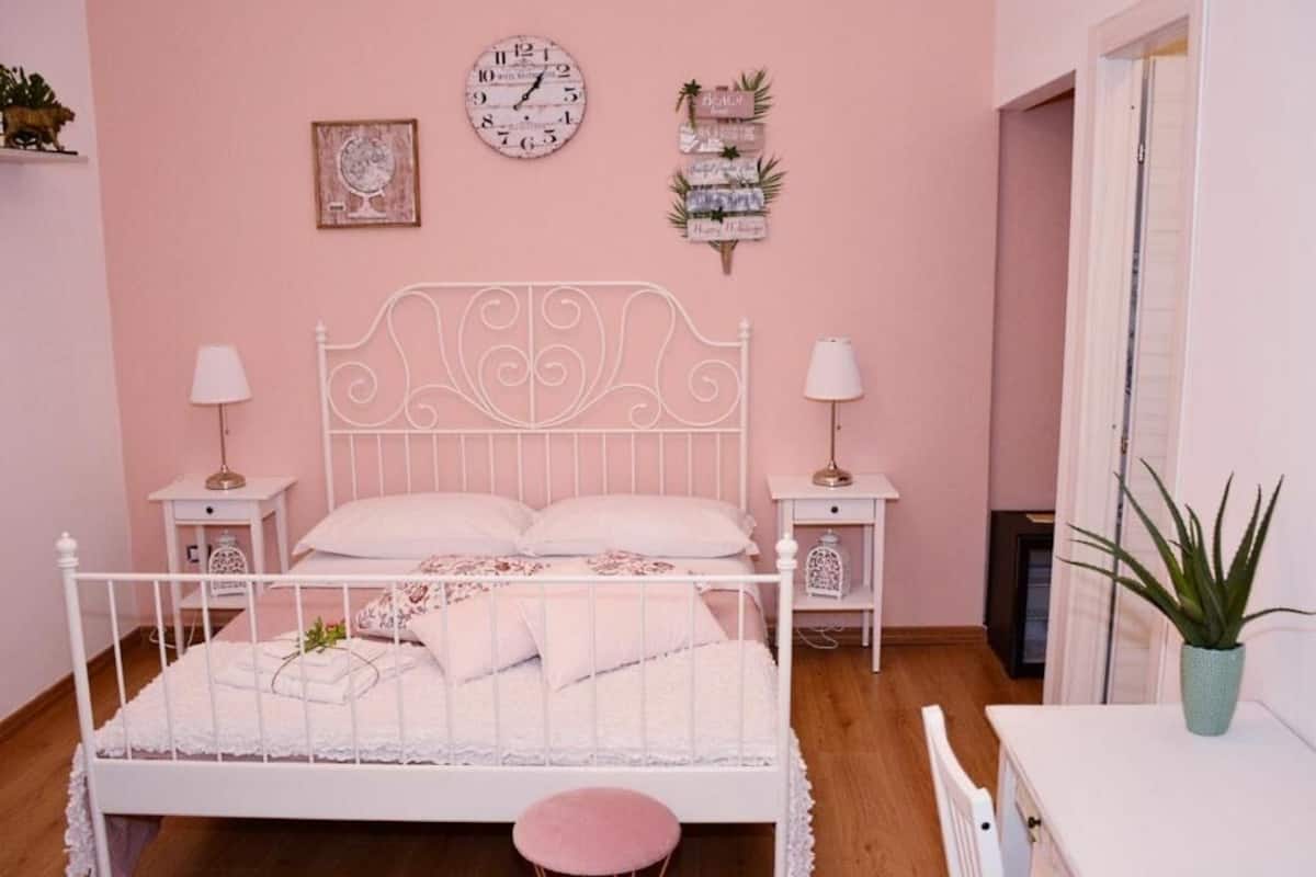 Un dormitorio con estructura de cama metálica blanca, dos mesitas de noche con lámparas, un reloj de pared redondo y un pequeño estante con adornos.