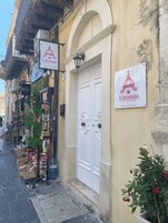 Una calle estrecha con un edificio que tiene una puerta blanca y un letrero que indica 'Lúrooms Guesthouse in Noto'.