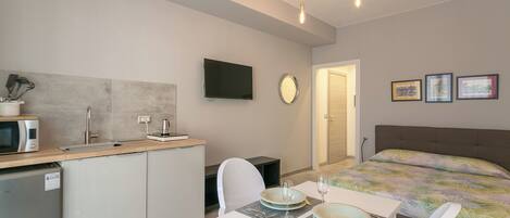 A compact hotel room with a kitchenette, a bed with a floral bedspread, a dining area with a table set for two, and a wall-mounted TV.