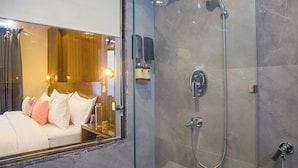Casa de banho moderna com cabine de duche em vidro, sanita à vista e uma cama com mesa de cabeceira no quarto adjacente.