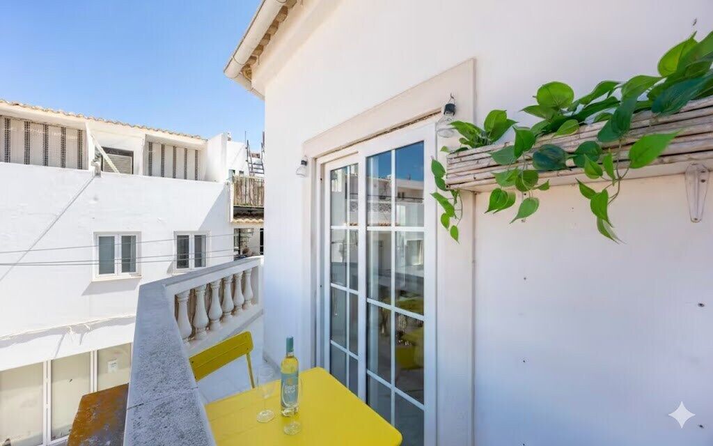 A balcony with a yellow table and a bottle, a white wall with a window, and a plant.