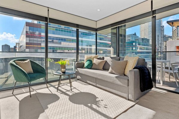Un salon moderne avec un canapé, un fauteuil et une petite table, de grandes fenêtres donnant sur la ville et un tapis à motifs.