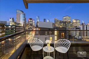Un balcon avec deux chaises blanches et une petite table, d’où l’on peut admirer la vue sur les toits de la ville au crépuscule.