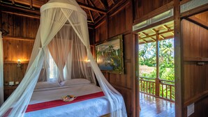 A wooden room with a bed under a canopy, a painting on the wall, and a view of greenery outside. - The Dukuh Retreat (Tabanan)