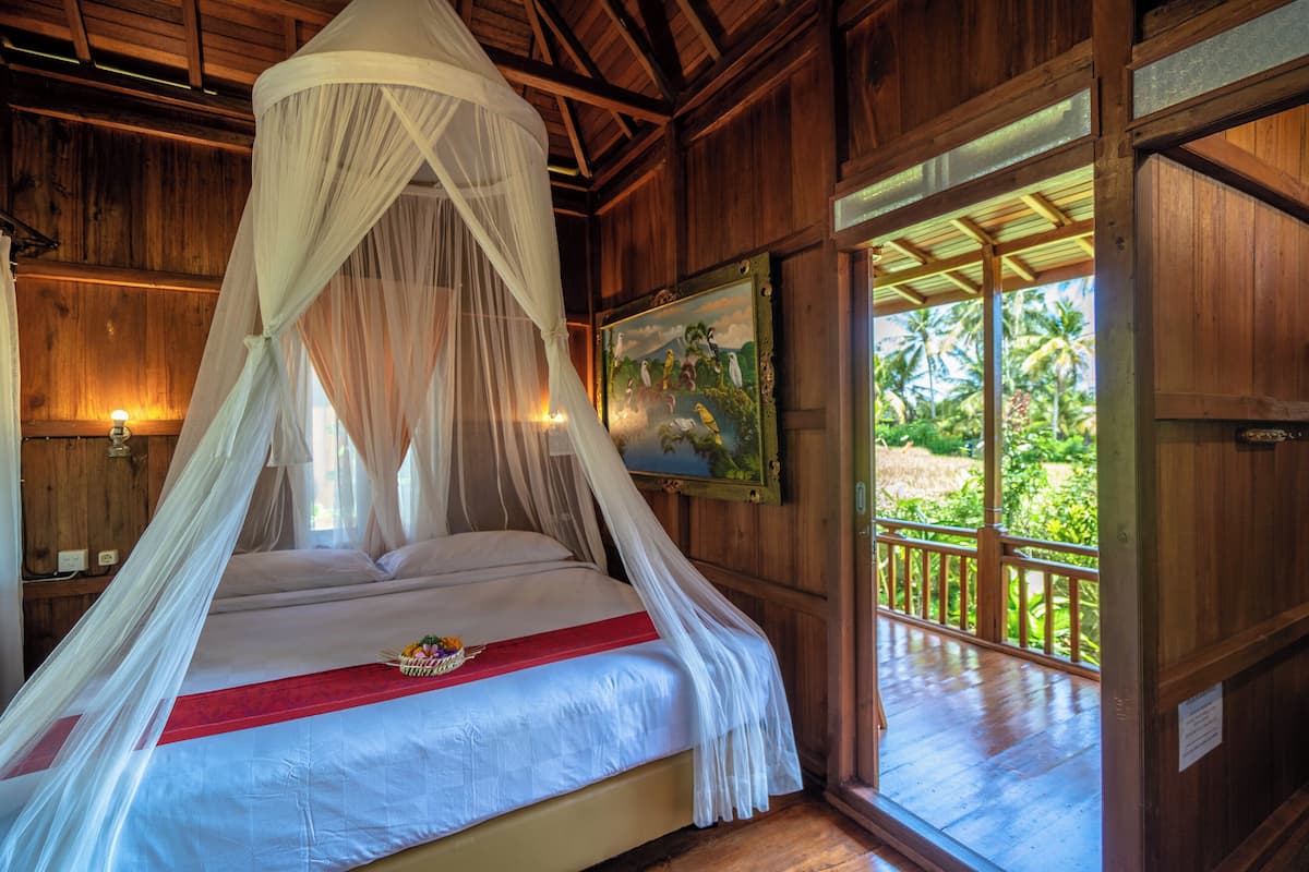 A wooden room with a bed under a canopy, a painting on the wall, and a view of greenery outside.