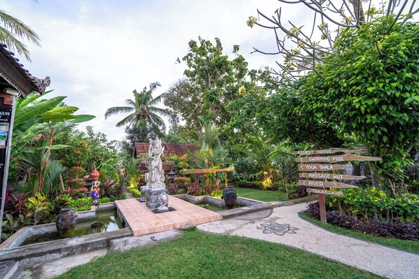 A garden with a statue, a pond, and a signpost with directions to amenities like a guest house, swimming pool, yoga shala, massage hut, and river. - The Dukuh Retreat (Tabanan)