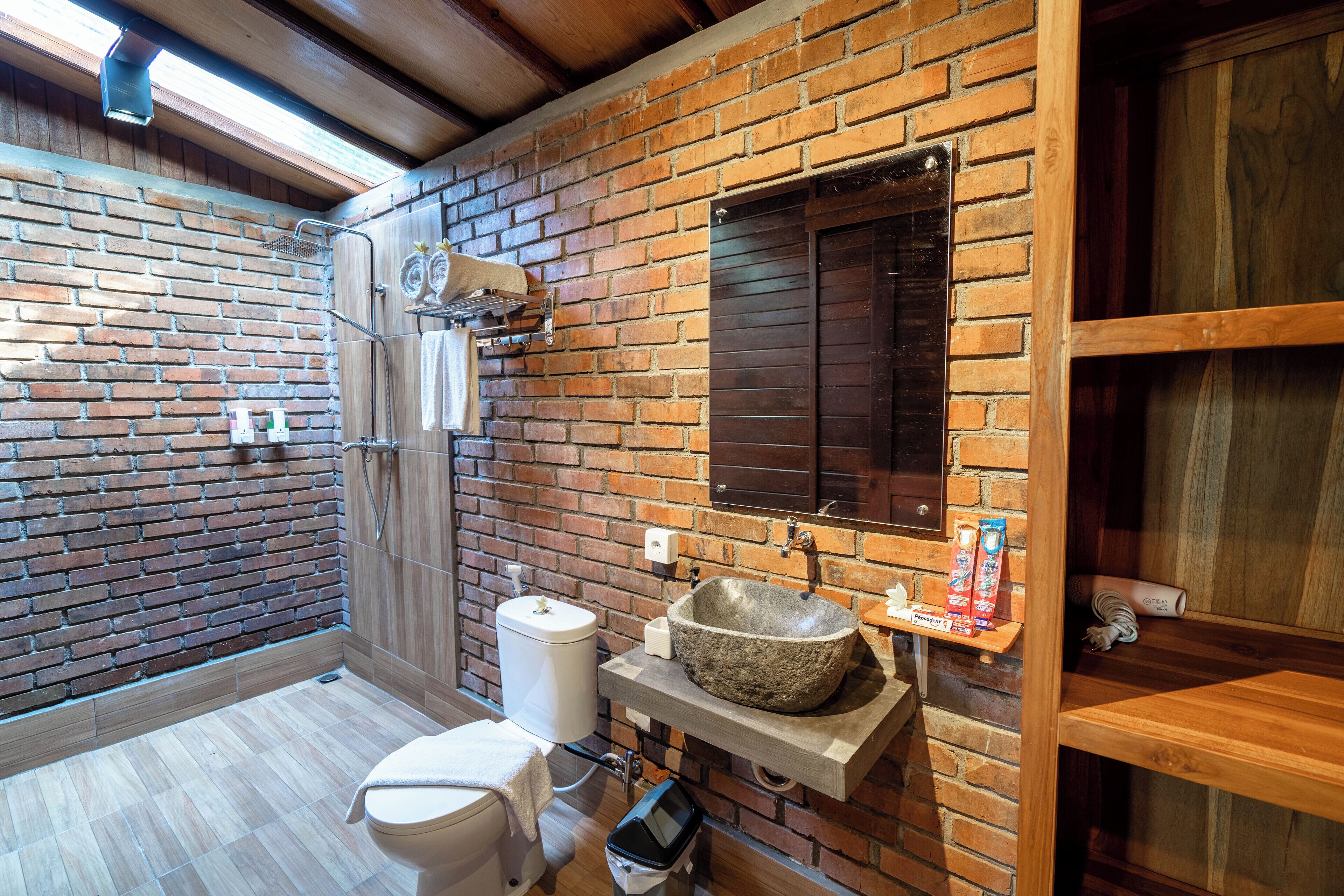 Un baño con un lavabo de piedra, un inodoro, una ducha y un estante de madera con artículos de aseo.