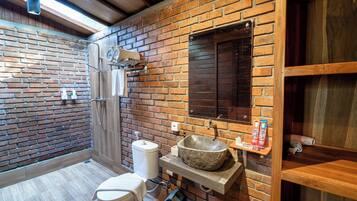 Un baño con un lavabo de piedra, un inodoro, una ducha y un estante de madera con artículos de aseo.