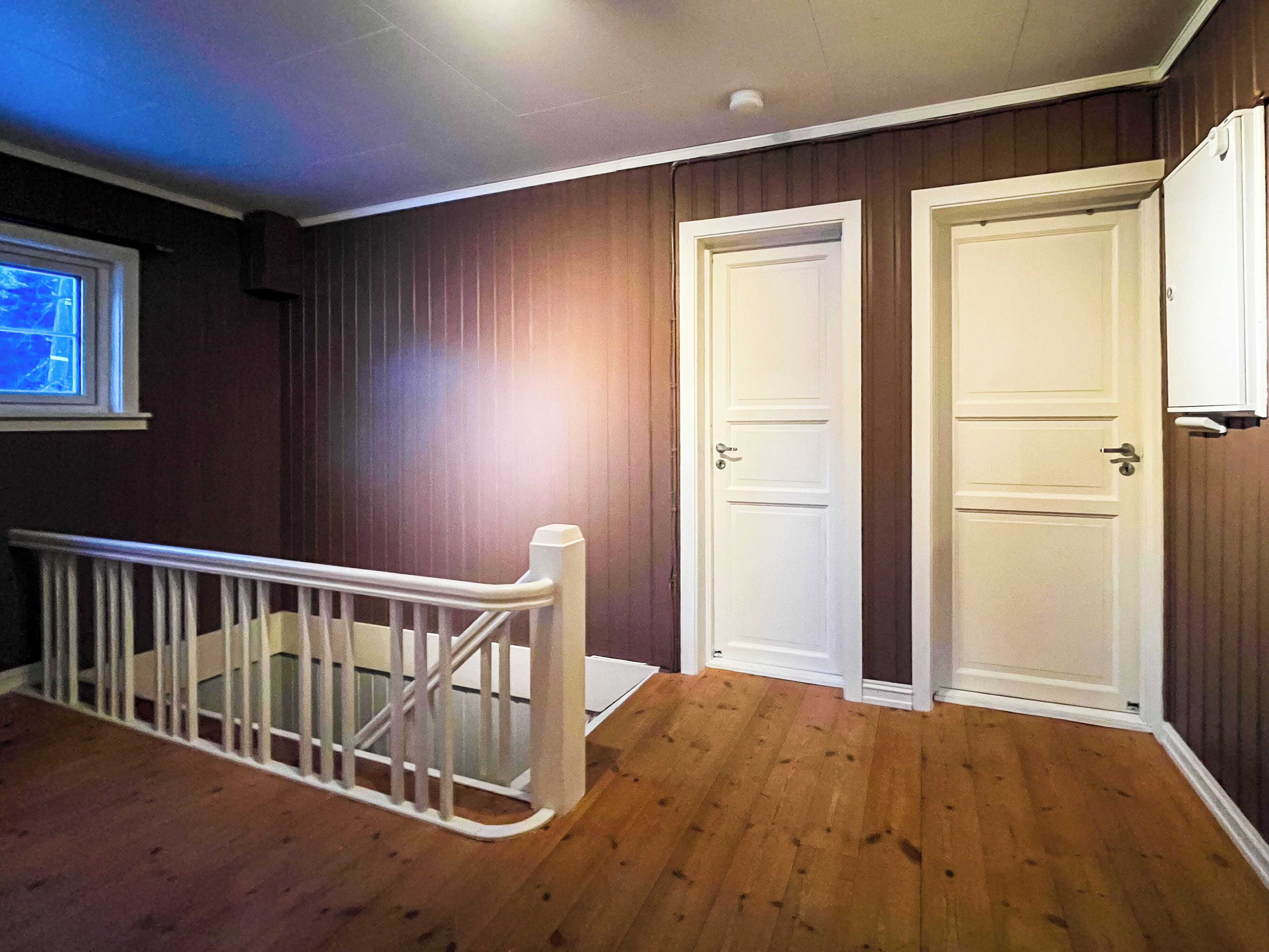 Une pièce avec un plancher en bois, deux portes blanches, une rambarde blanche et une fenêtre avec des stores.
