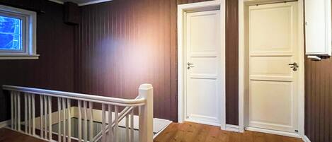 Une pièce avec un plancher en bois, deux portes blanches, une rambarde blanche et une fenêtre avec des stores.